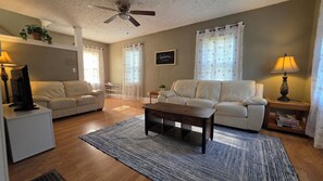 A living room with a sofa, a coffee table, a TV on a stand, a ceiling fan, and a welcome sign.