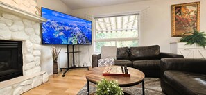 A living room with a fireplace, a flat-screen TV, a leather sofa, a coffee table with a chess set, and a painting on the wall. - Cozy 4BR Retreat | Entire House | King Bed | Airport | Ring Road | Games | Banff (Calgary)