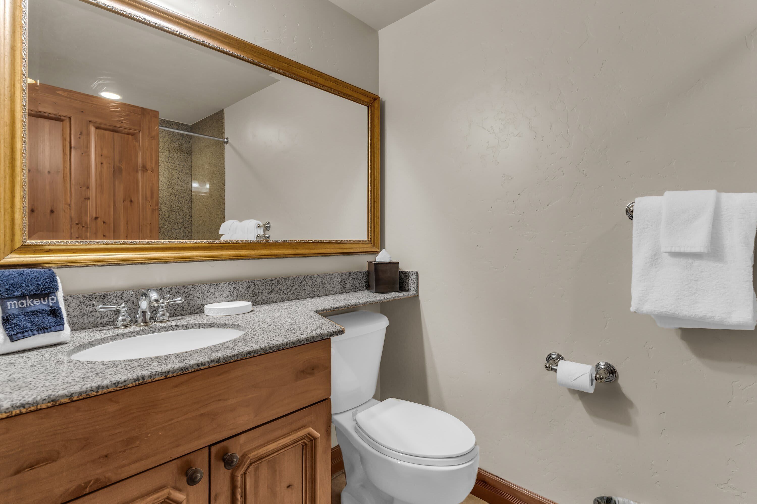 A bathroom with a granite countertop sink, wooden vanity, a white toilet, and a large mirror with a gold frame.
