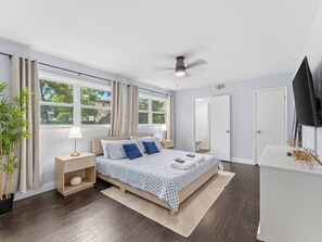 A modern bedroom with a bed, bedside tables, a ceiling fan, a television, and a large window with curtains. - Modern Gulf Coast Getaway| Near Clearwater and Indian Rocks Beach (Largo)