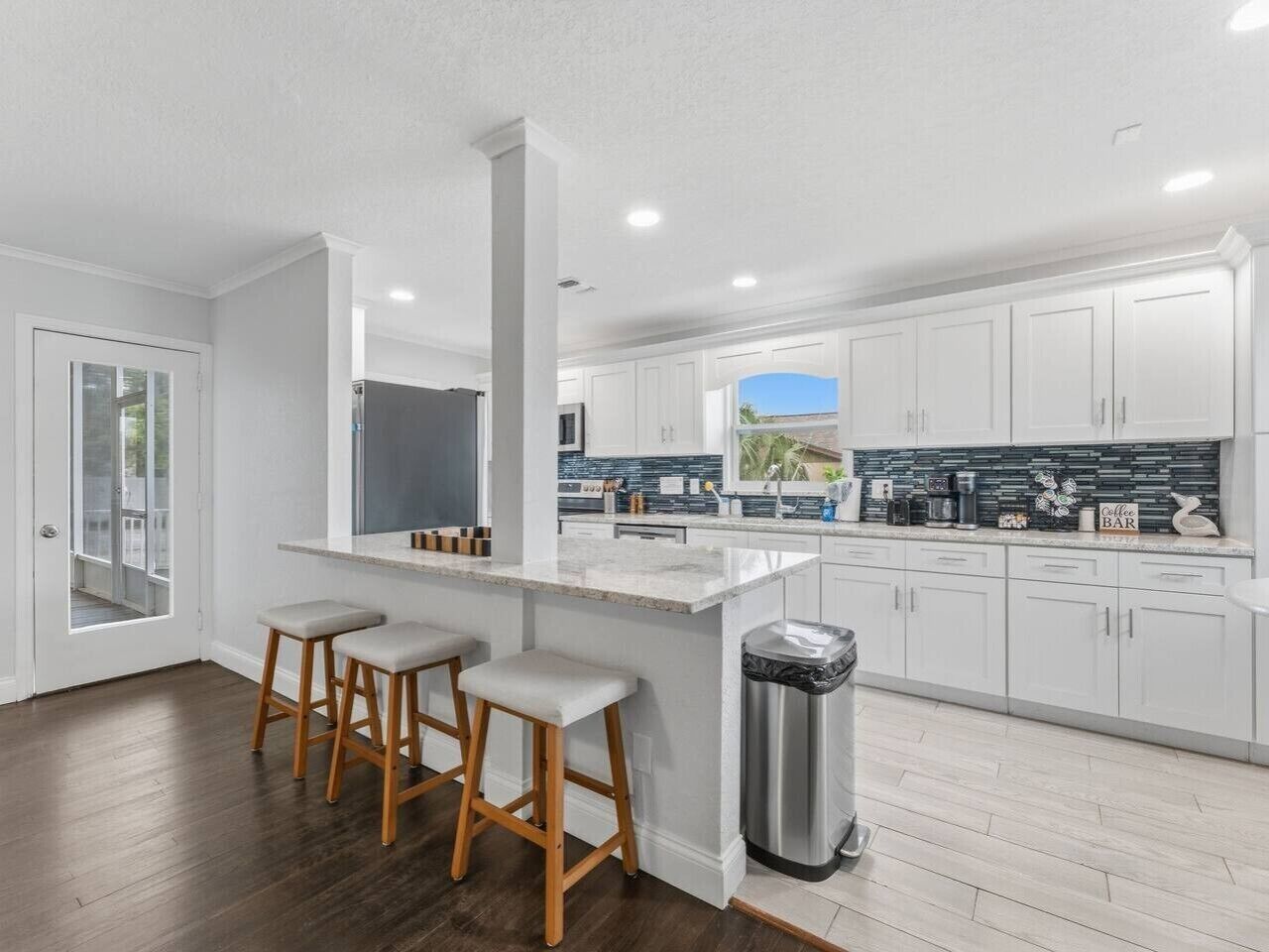 A modern kitchen with white cabinets, a central island with stools, and a view through a glass door.