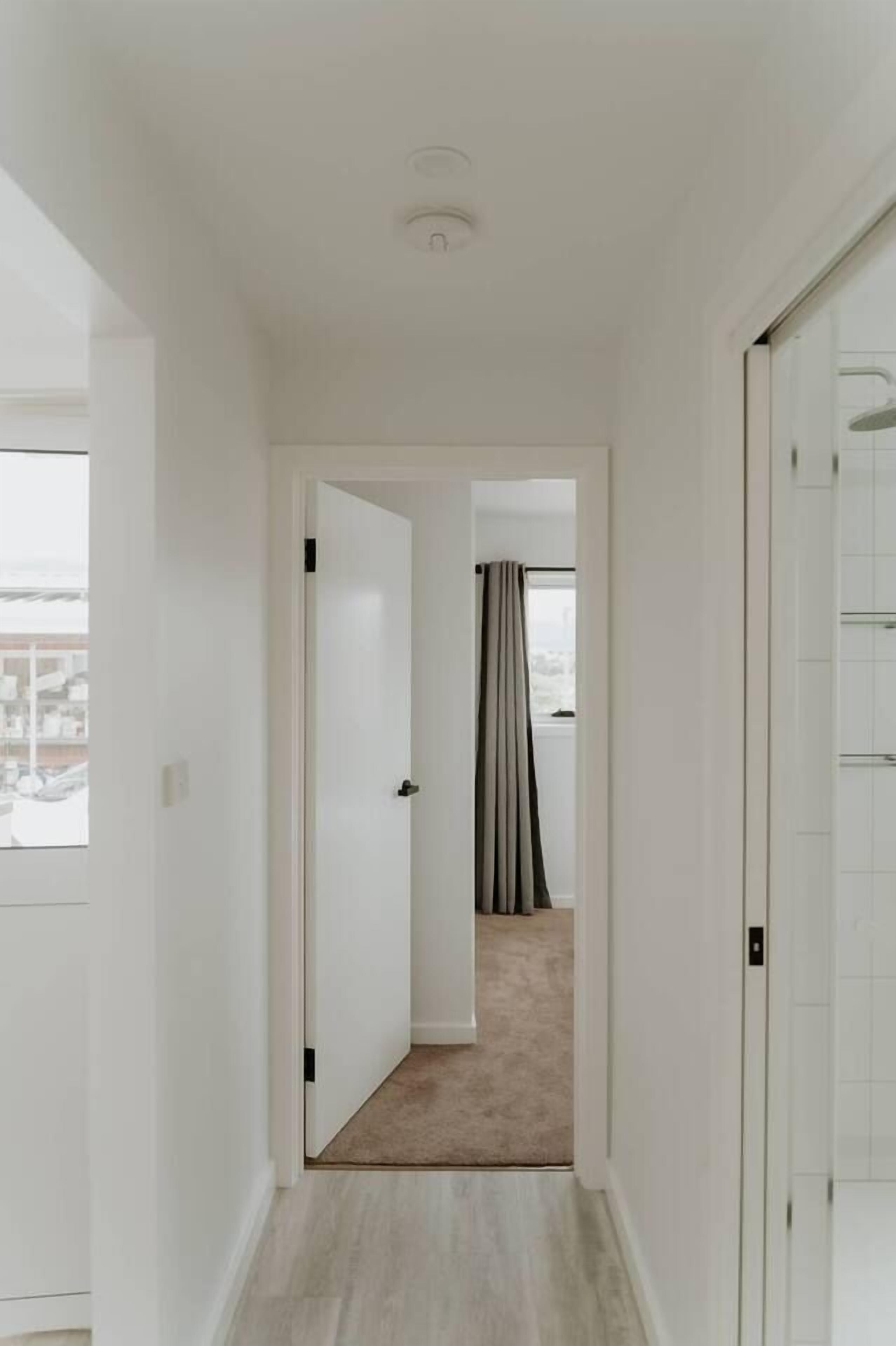 A narrow hallway with white walls and a carpeted floor leading to a room with a window and curtains.