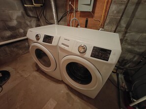 Eine Samsung Waschmaschine und Trockner mit Frontlader in einem Waschraum, wobei Sanitär- und Elektroanschlüsse sichtbar sind.