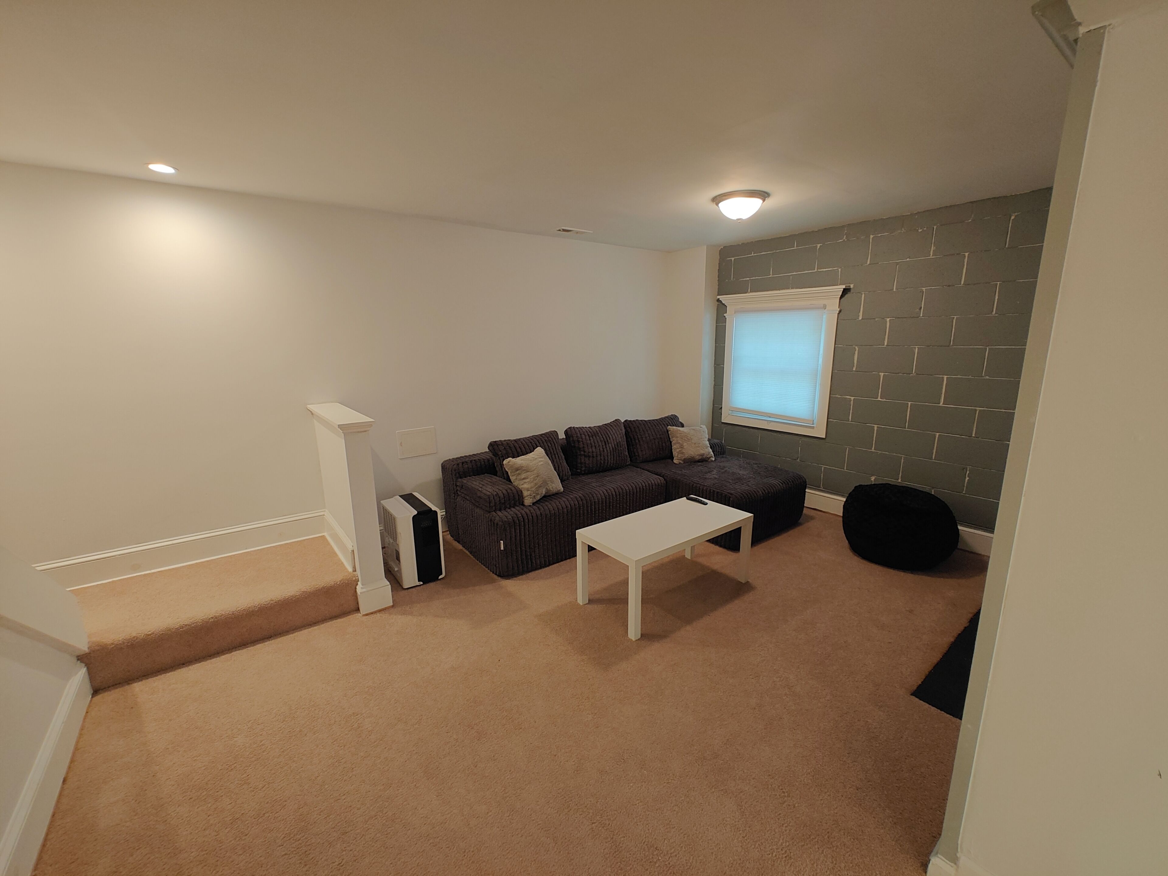 A room with a brown carpet, a white table, a brown sofa with cushions, a white radiator, and a window with blinds.