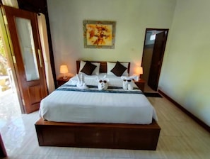 A neatly made bed with pillows and a blanket, a nightstand with a lamp, a painting on the wall, and a door to the left.