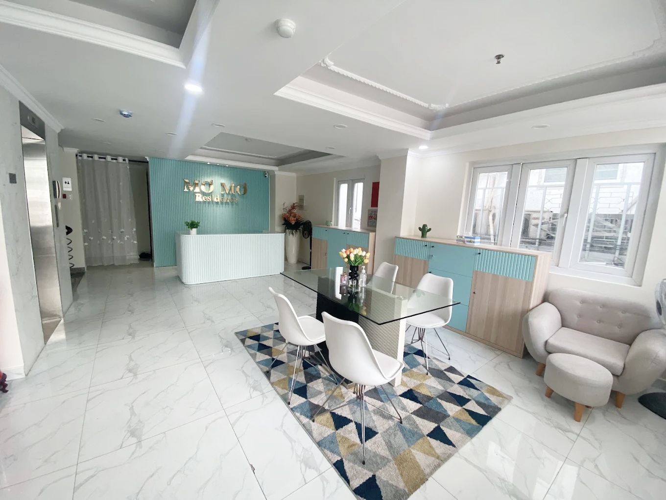 A modern hotel reception area with a glass desk, white chairs, and a geometric patterned rug.