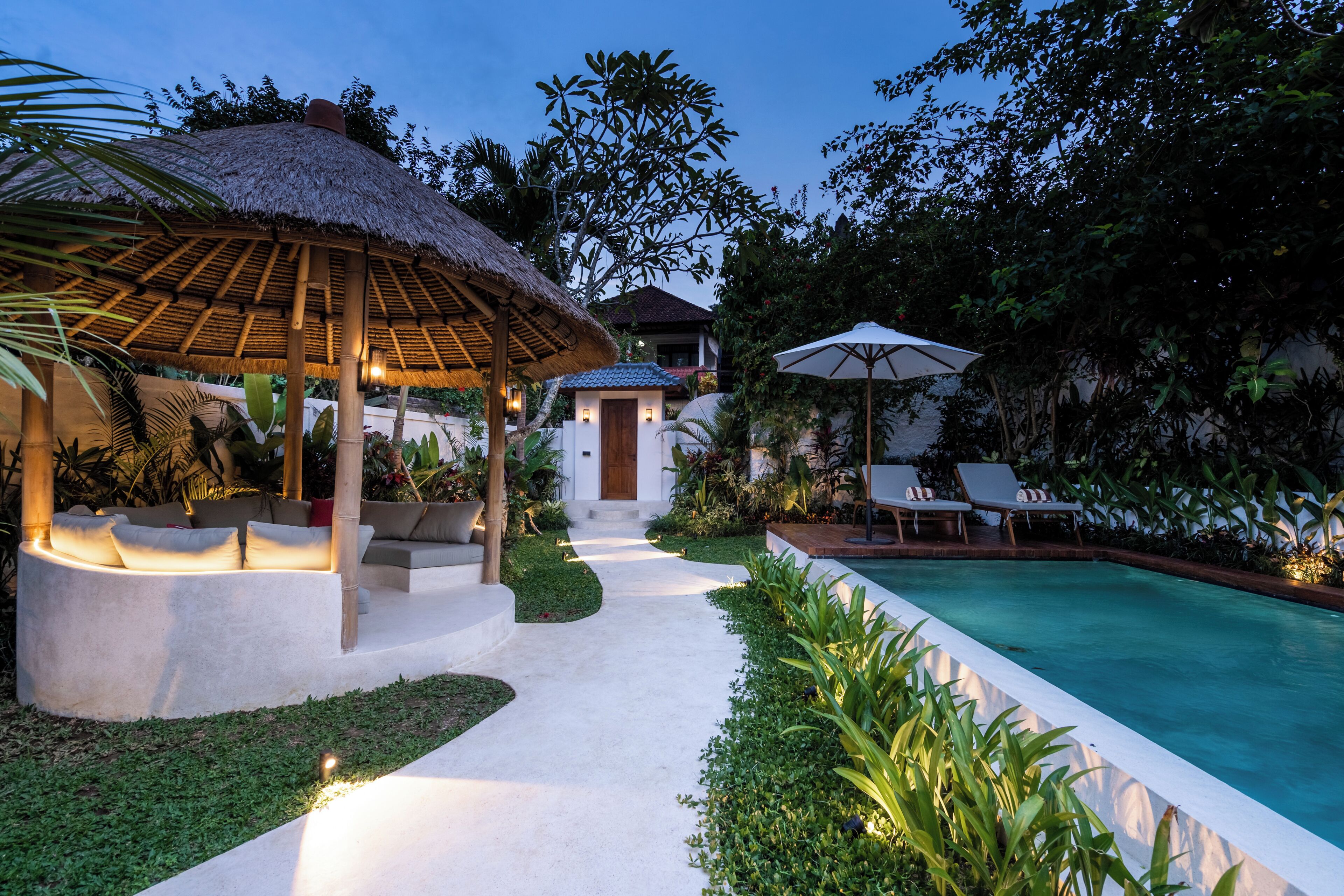 A poolside area with a thatched gazebo, lounge chairs, and a pathway leading to a building.