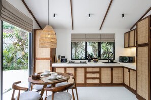 A modern kitchen with a dining area, featuring a large window, a hanging light fixture, and a built-in stove. - Capri Villa by DH (Gianyar)