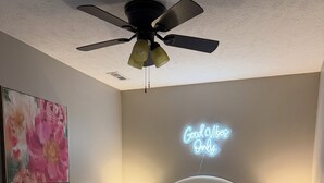 A bedroom with a bed, two bedside tables, a ceiling fan, a neon sign, and a floral painting on the wall.