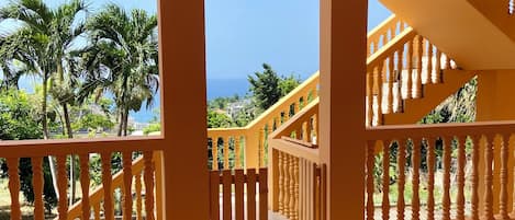 A spacious indoor area with a tiled floor, a staircase with a balustrade, and a view of palm trees and buildings through a window.