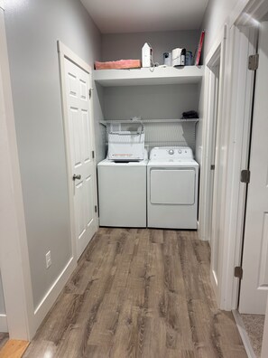 A laundry room with two white washers and dryers, wooden flooring, and a shelf with cleaning supplies. - Fabulous 2-bedroom house with AC in charming Sandusky (Sandusky)