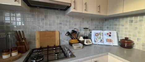 Una cocina con estufa a gas, tabla de cortar de madera, licuadora y libro de cocina sobre el mostrador.
