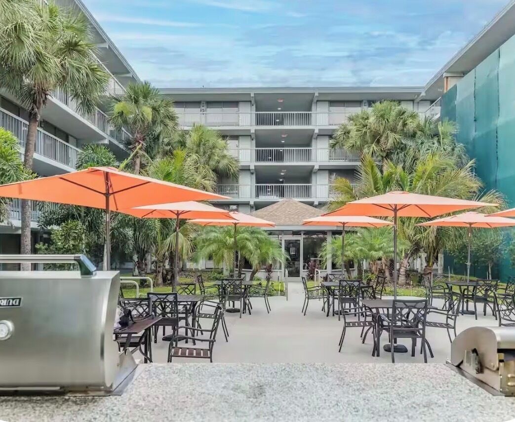 An outdoor patio area with tables and chairs, surrounded by palm trees and covered by orange umbrellas.