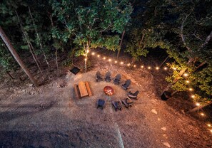 An outdoor seating area with a fire pit, surrounded by chairs and a bench, set in a forested area with string lights. - The Revel | 24-Guest Luxury Cabin + Views (Broken Bow)