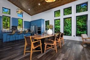 A modern kitchen with blue cabinets, a dining area with wooden chairs, and large windows offering a view of greenery. - The Revel | 24-Guest Luxury Cabin + Views (Broken Bow)