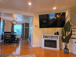 A living room with a flat-screen TV mounted on the wall, a fireplace, a dining area with a table and chairs, and a staircase. - Luxury Cozy Stay-Near Six Flags, Navy Base (Gurnee)