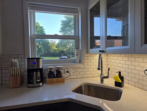 A kitchen with a stainless steel sink, a coffee maker, and a window with a view of trees. - Luxury Cozy Stay-Near Six Flags, Navy Base (Gurnee)