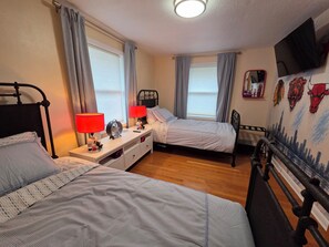 Ein Schlafzimmer mit zwei Betten, einem Nachttisch mit Lampen, einem Ventilator und einer Wand mit Stadtbild-Wandmalerei.
