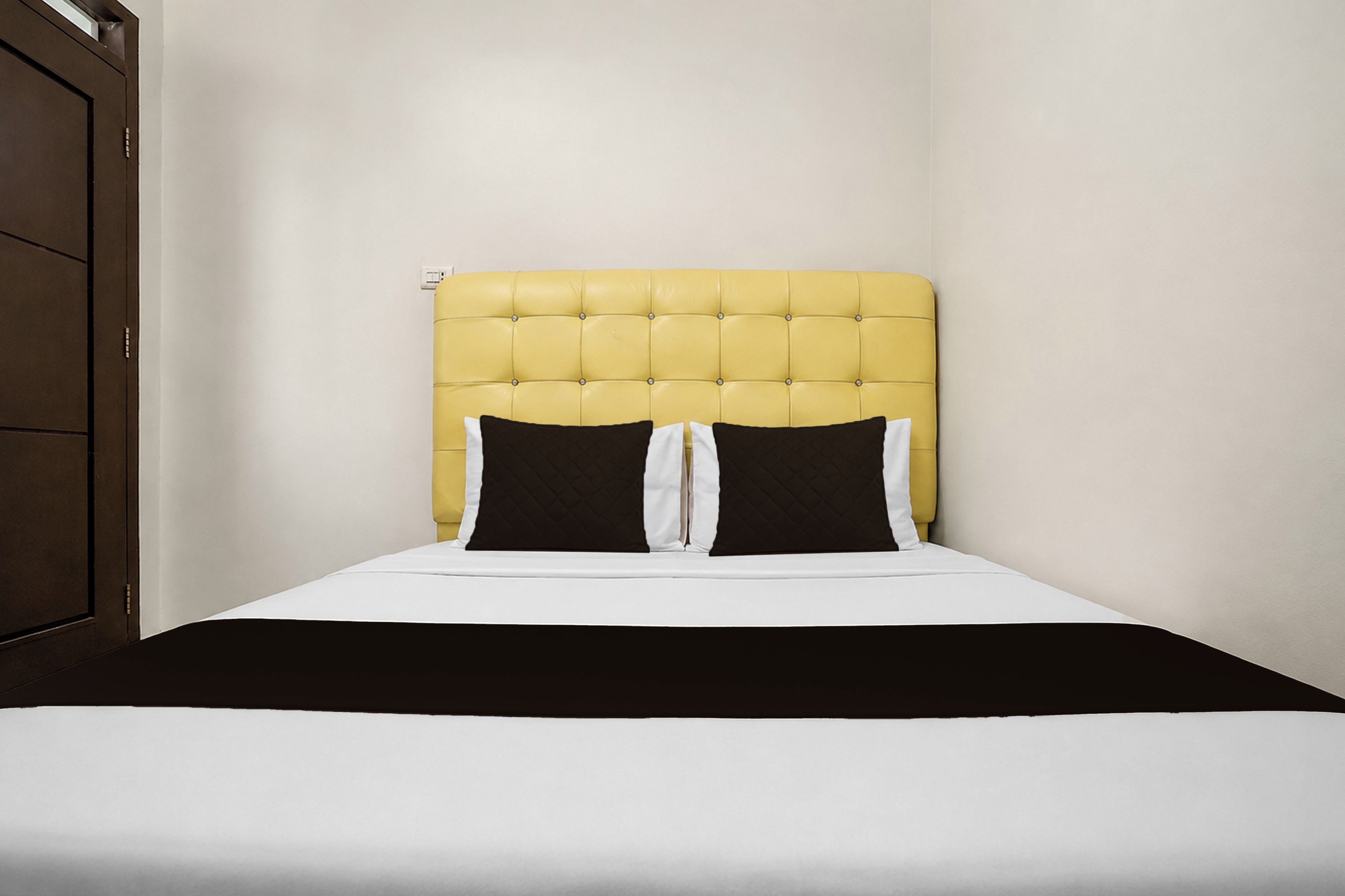 Un lit simple avec une tête de lit rembourrée de couleur jaune, une literie blanche et deux oreillers noirs.