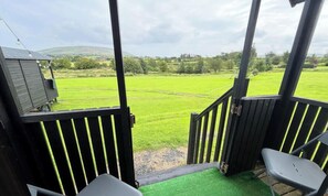 Ein überdachter Außenbereich mit Blick auf ein grünes Feld, ein kleines Gebäude und einen Stuhl.