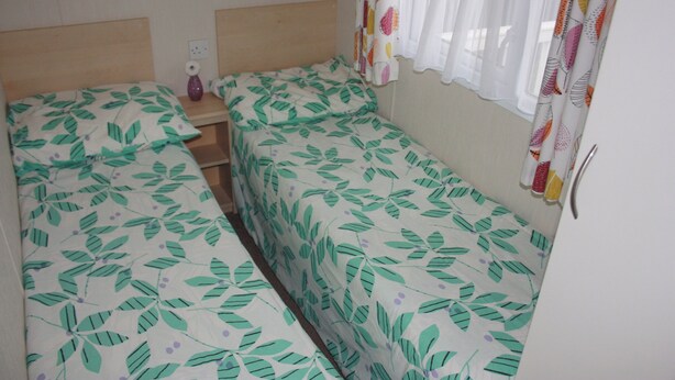 A small, single-room accommodation with two single beds, a bedside table, and a window with curtains. - 10 Berth Caravan Butlins Skegness 2 showers pets (Skegness)