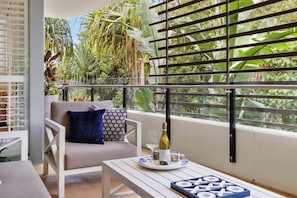 A balcony with a white chair, a table with a wine bottle, and a view of lush greenery. - Luxury 3-bedroom Penthouse Apartment (Casuarina)