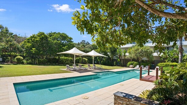 Una piscina rectangular con sillones de descanso y sombrillas, rodeada de un jardín ajardinado y un edificio al fondo.