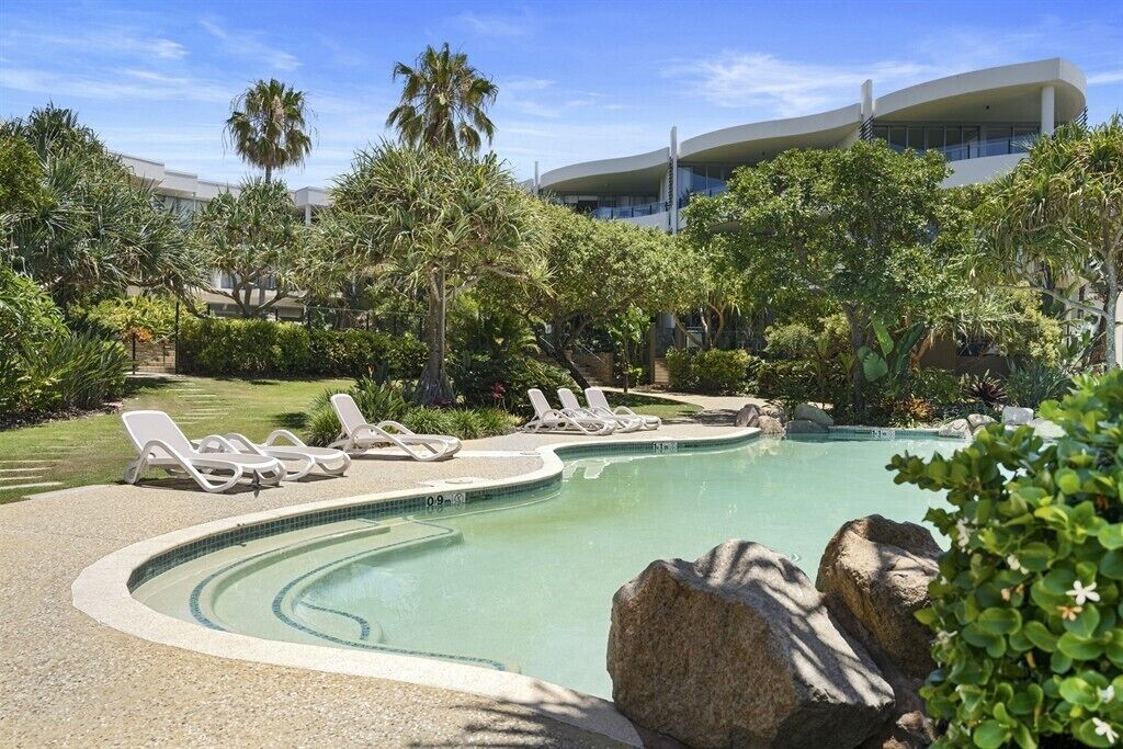 Une piscine entourée de chaises longues, de plantes tropicales et, au loin, d’un bâtiment moderne.