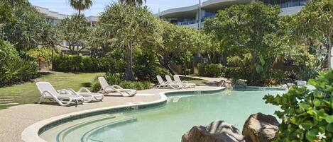 Une piscine entourée de chaises longues, de plantes tropicales et, au loin, d’un bâtiment moderne.