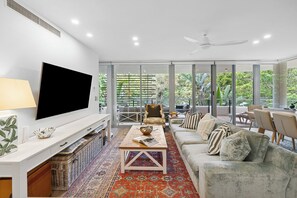 Ein modernes Wohnzimmer mit Flachbildfernseher, einer Couch, einem Couchtisch und einem großen Fenster mit Blick ins Grüne.