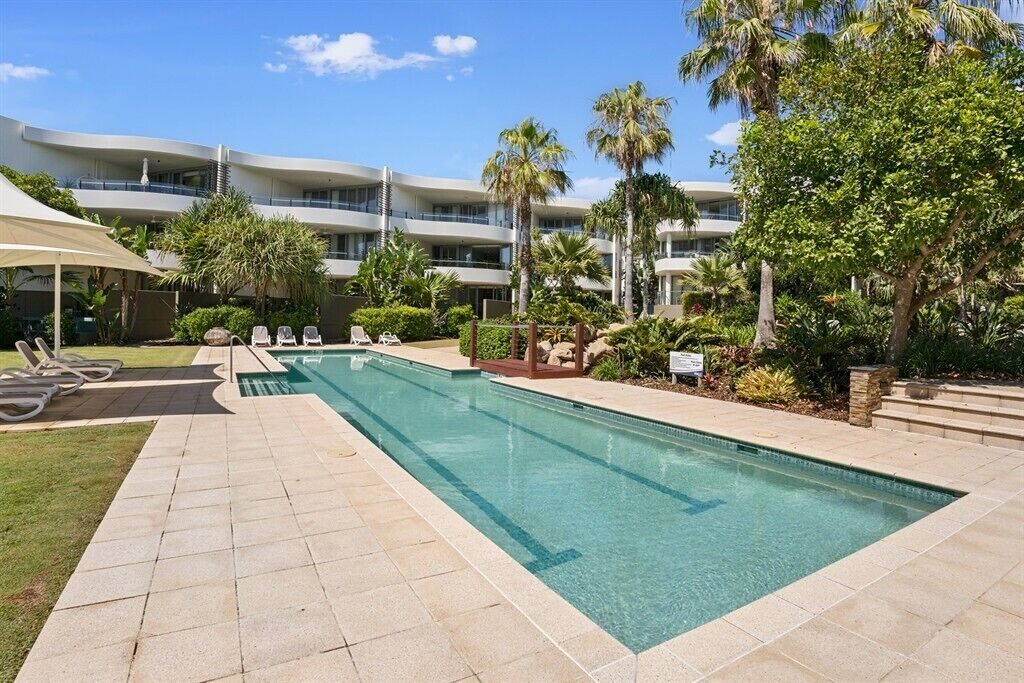 A swimming pool with lounge chairs, palm trees, and a multi-story building in the background.