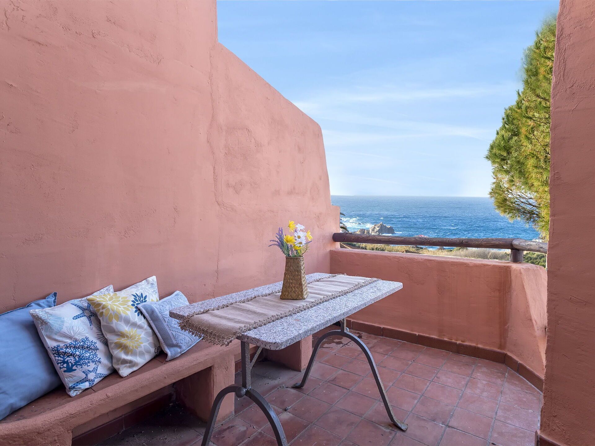 A balcony with a table, bench, and pillows, overlooking the sea.