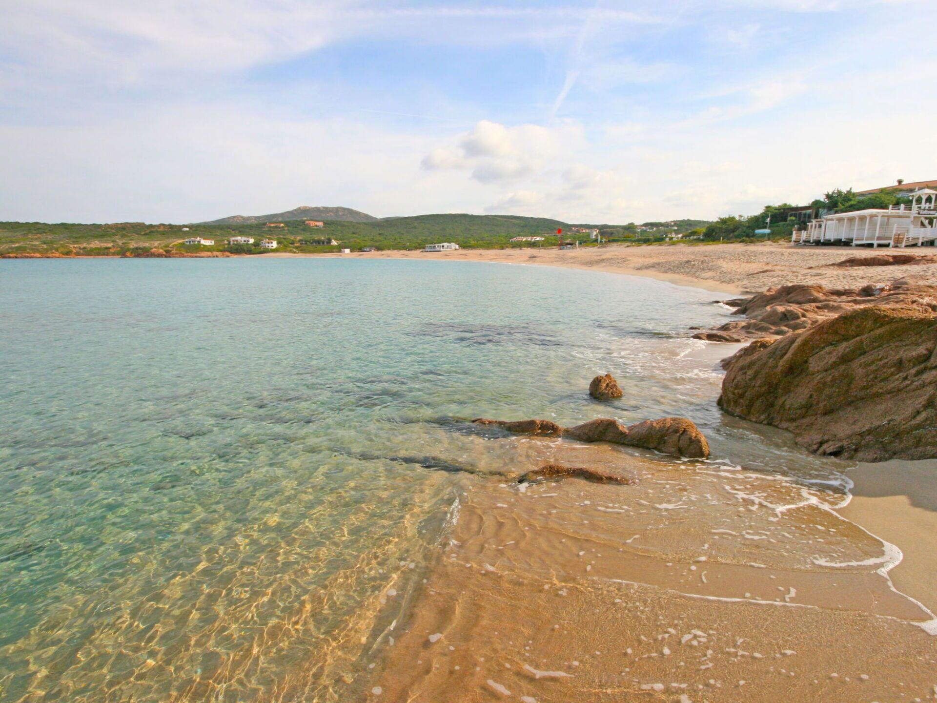 A beach with clear water, rocky shoreline, and buildings in the background.