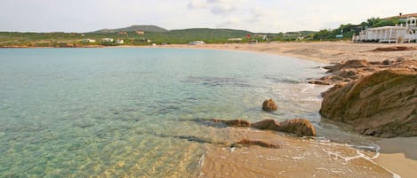 Una playa con agua cristalina, costa rocosa y edificios al fondo.
