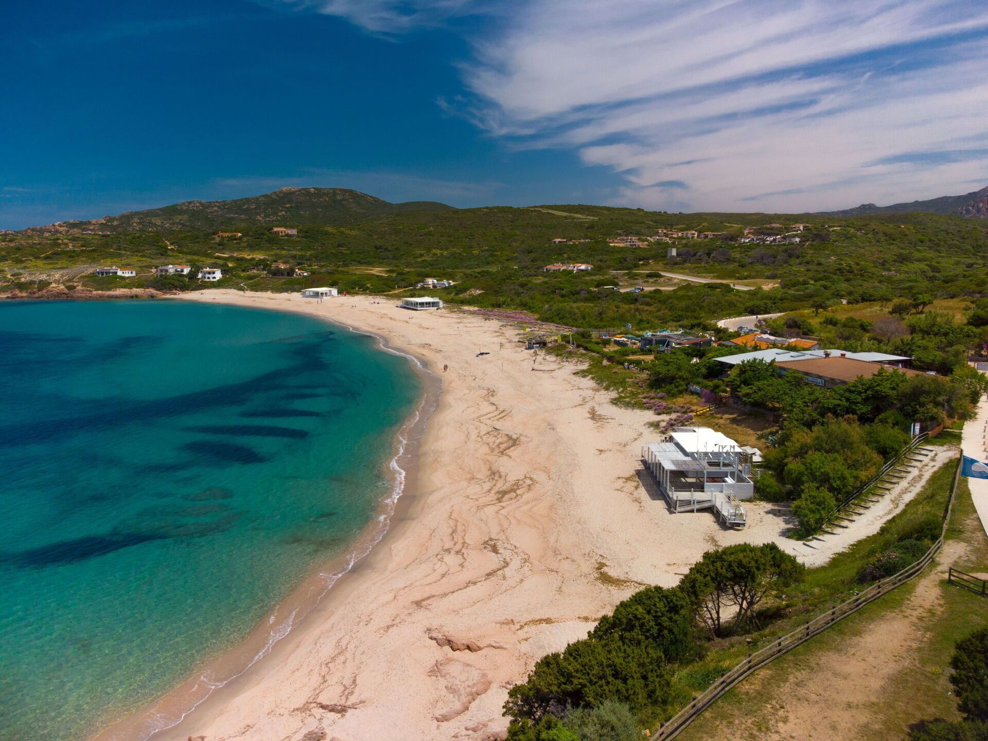 Vista aérea de una zona costera con playa de arena, aguas azules cristalinas y edificios dispersos.
