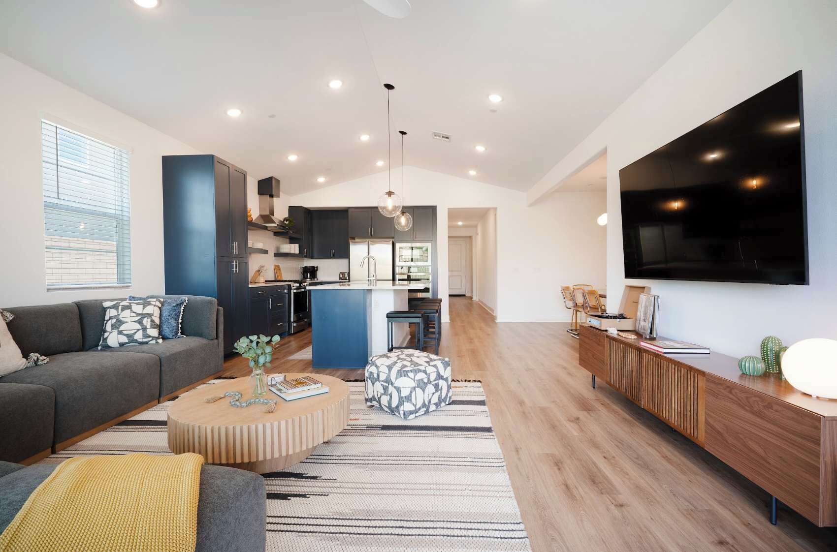 Una sala de estar moderna con un sofá, un reposapiés y una mesa de centro, una cocina al fondo y un televisor montado en la pared.