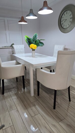 A dining area with a white table, two chairs, pendant lights, a clock on the wall, and a potted plant with yellow flowers. - King size bed ensuite bathroom wifi near university hospital Birmingham (Birmingham)
