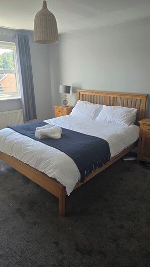 Een net opgemaakt bed met witte lakens en een donkerblauwe plaid, een houten hoofdbord, een bedlamp en een raam met gordijnen.