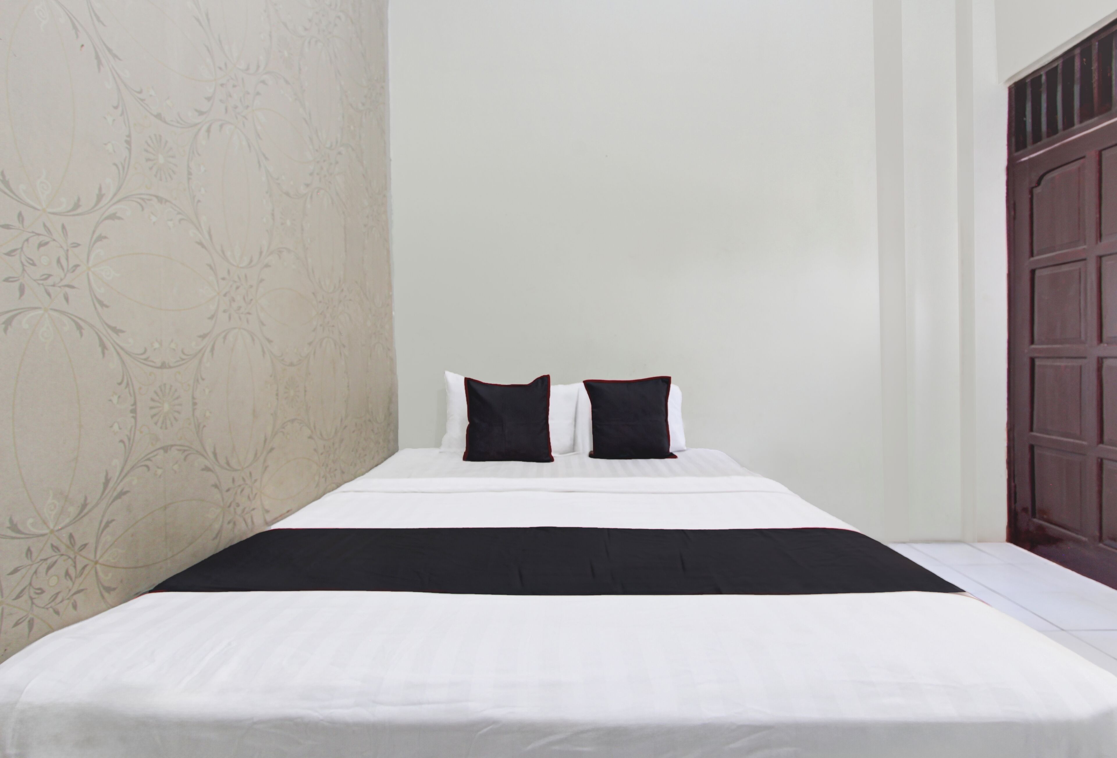 A neatly made bed with white bedding and two black pillows, a dark throw blanket, and a floral wallpaper pattern on the wall.