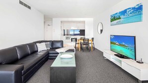 A modern living room with a black leather sofa, a glass coffee table, a dining area with a wooden table and chairs, a kitchen in the background, and a large TV on the right. - Circle on Cavill Ocean View (Surfers Paradise)