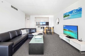 A modern living room with a black leather sofa, a glass coffee table, a dining area with a wooden table and chairs, a kitchen in the background, and a large TV on the right. - Circle on Cavill Ocean View (Surfers Paradise)