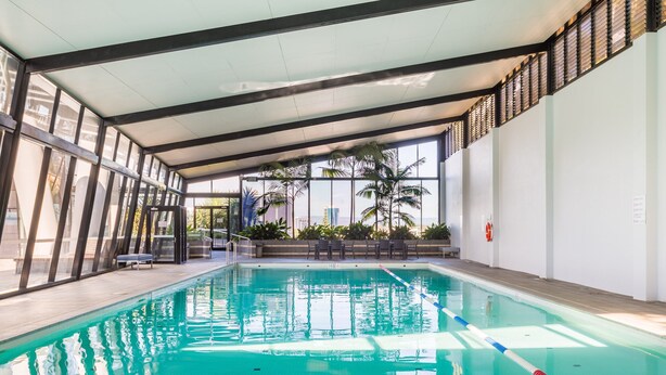 An indoor swimming pool with clear water, surrounded by glass walls and a view of palm trees and buildings. - Circle on Cavill Ocean View (Surfers Paradise)