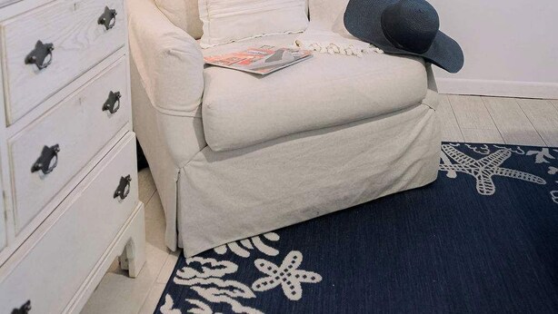Una habitación con un sillón blanco, una mesita de noche con una lámpara, una obra de arte enmarcada de un pájaro y una alfombra con estampado de estrella de mar.