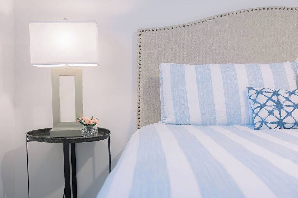A bed with striped bedding, a bedside lamp, and a small table with a vase of flowers.
