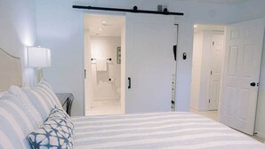 Un dormitorio con una cama, una mesita de noche, una lámpara y una puerta que da al baño.