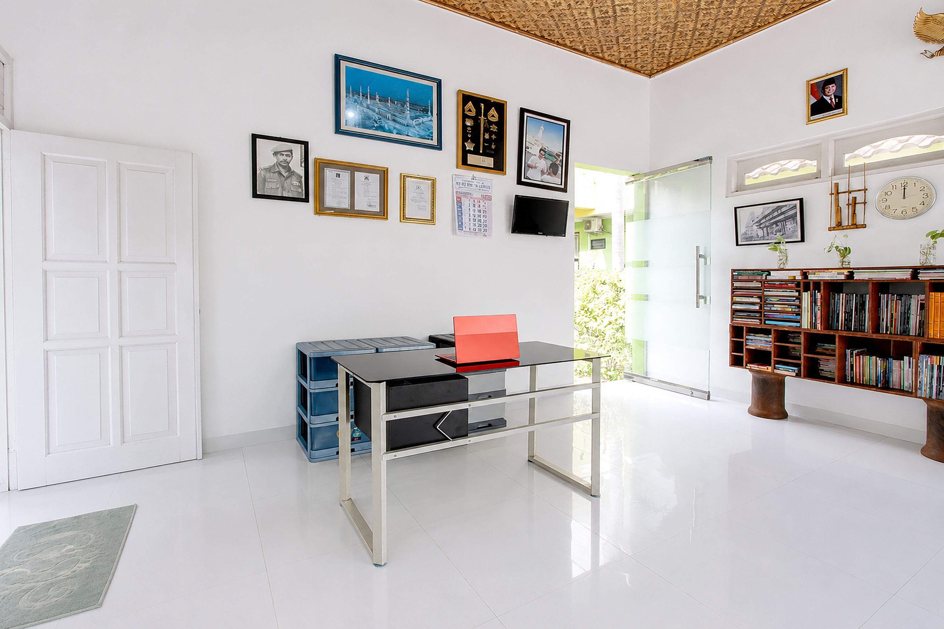A room with a white door, a desk with a laptop, a bookshelf, and various framed items on the wall.