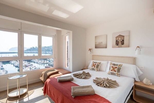 A bedroom with a large bed, a side table, a round table, and a view of a marina through large windows. - Geure portue etxea, wonderful sunrise (Bermeo)