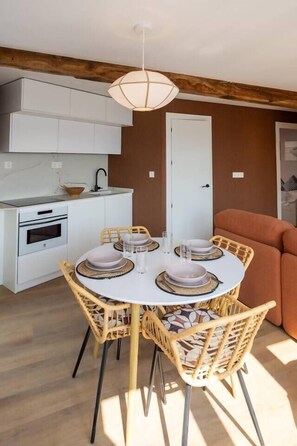 A compact kitchen with white cabinets, a built-in oven, and a dining area with a round table and wicker chairs. - Geure portue etxea, wonderful sunrise (Bermeo)