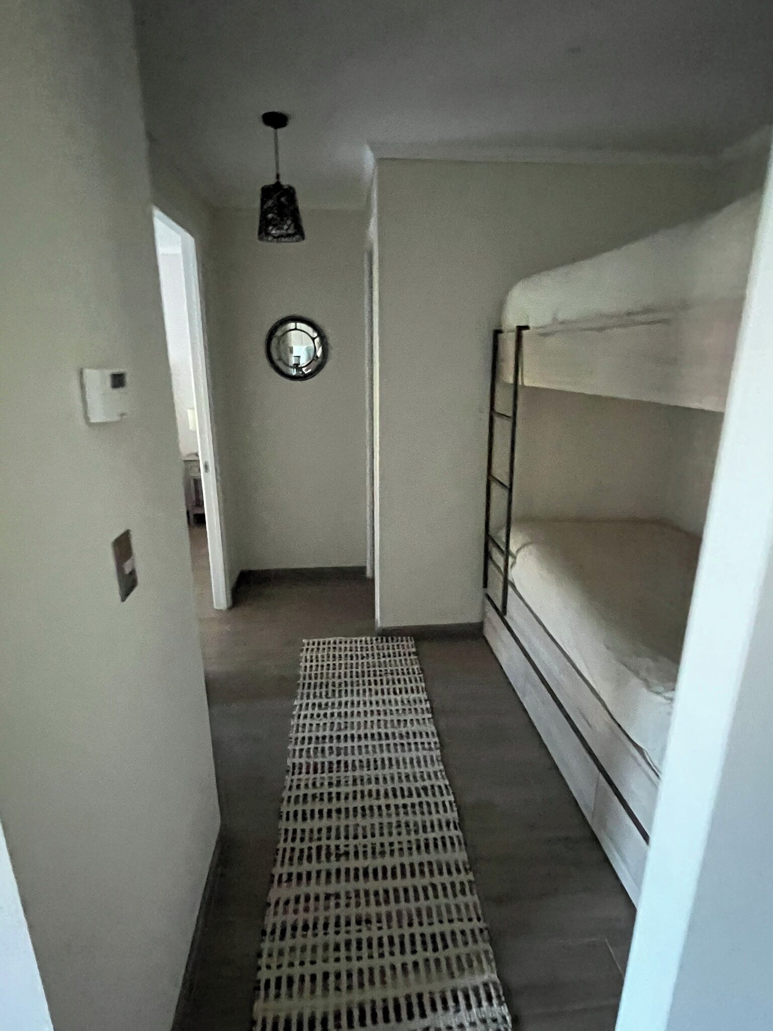 A narrow hallway with a bunk bed on the right, a door at the end, and a small window with a circular decoration on the wall.