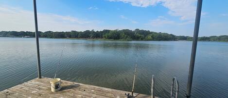 A wooden dock with fishing rods and a bucket, overlooking a calm lake with a forested shoreline.